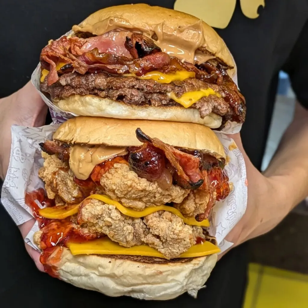Two Sneaky Burger stacks held up — a beef burger and a fried-chicken burger loaded with bacon and sauce