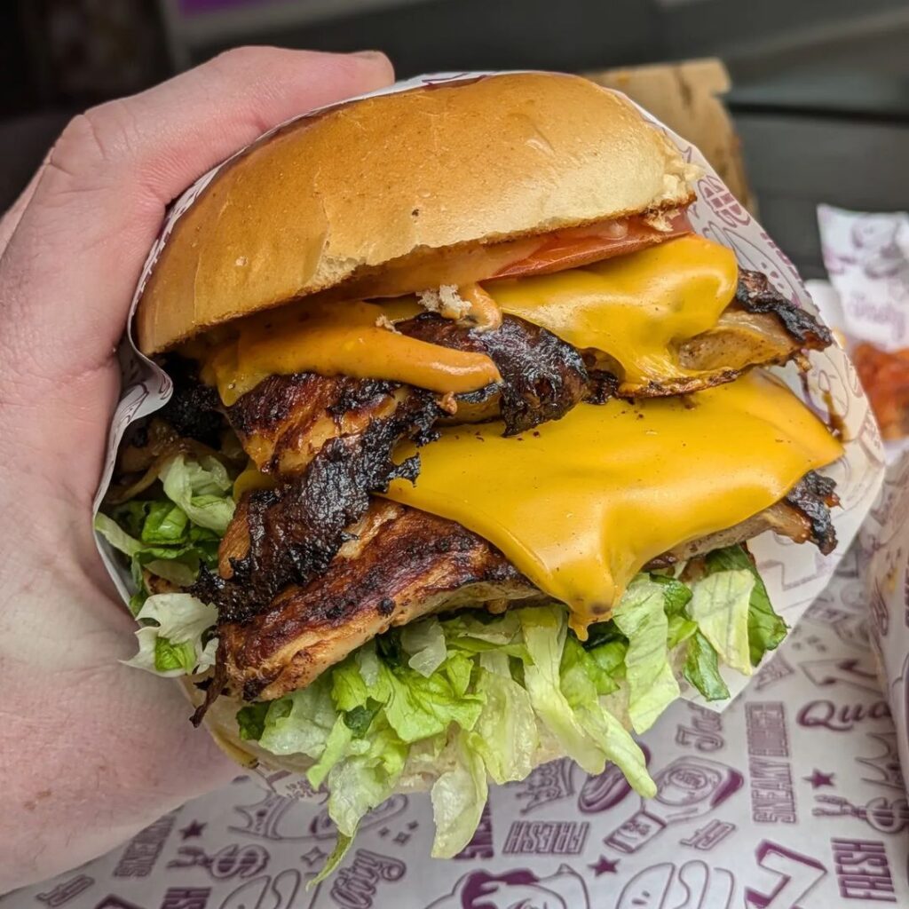 A hand holds a grilled chicken sandwich with two layers of cheddar cheese, shredded lettuce, and tomato, encased in a paper wrapper. The sandwich appears juicy and freshly made, with visible grill marks on the chicken. In the background, part of a tray and paper are visible.