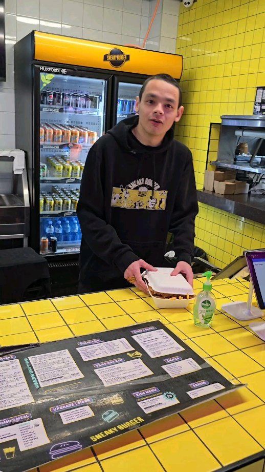 A person wearing a black hoodie stands behind a counter in a restaurant with vibrant yellow tiles. The counter has a menu and a hand sanitizer bottle. Refrigerated beverages are stocked in a fridge behind them. The person is holding a white box with food.